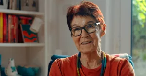 Ailbhe Smyth in a video for Belong To. She is on the right hand side of the frame, sitting and looking to someone behind the camera. There is a bookshelf on the left hand side of the frame.