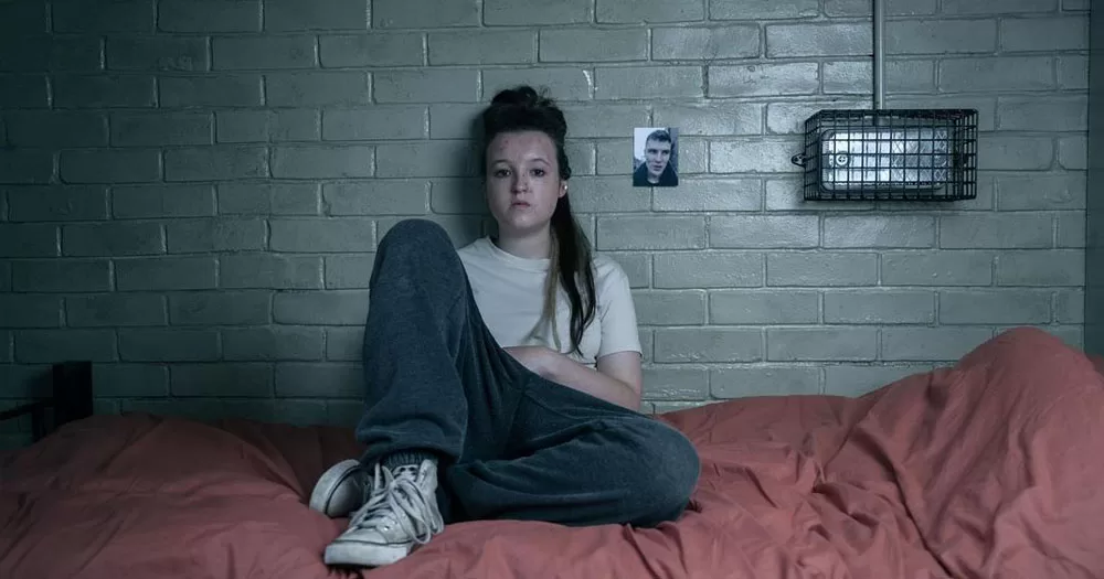 The image shows neurodivergent actor Bella Ramsey sitting on a bed in a prison cell. They are wearing jeans and a white tshirt and are leaning against a brick wall. They have long hair and red marks on their face.