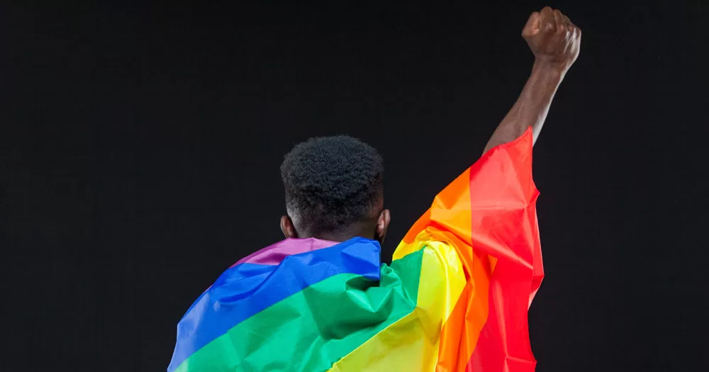 The image marks Black History Month. I shows the back of a person wrapped in a Pride flag in front of a black background. The person has short crew cut hair with tight curls on top. They have one arm raised in a fist.