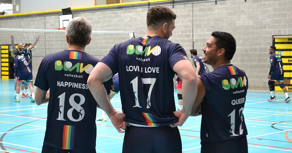 An image of the back of three Bravo Volleyball players. They are pictured from the waist up, and their navy jerseys with their numbers, club logos and slogans on them are visible. A volleyball court is also visible in the background.