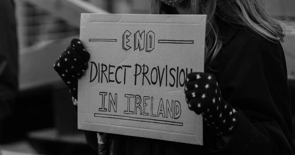 The Ombudsman for Children has issued a new report raising concerns over children in Direct Provision. The black and white image shows a person with long hair holding up a cardboard sige which reads "End Direct Provision in Ireland". The person is wearing black fleece gloves with white stars.