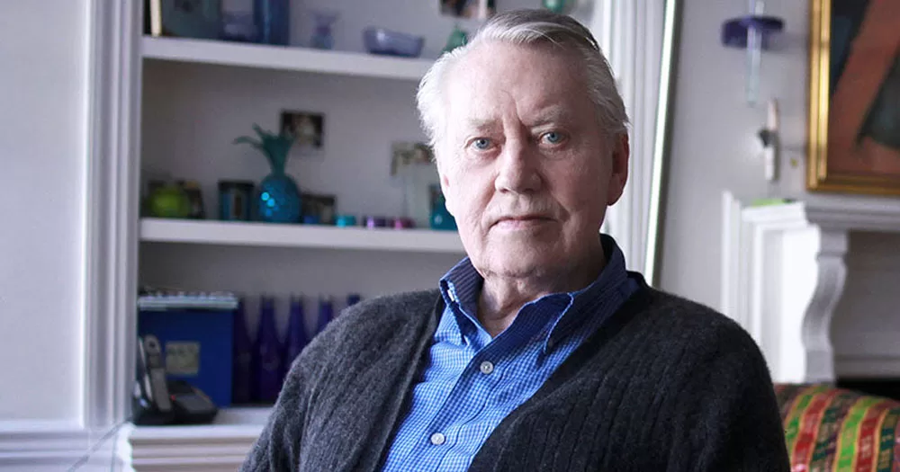 The image is a portrait of philanthropist, Chuck Feeney. He is wearing a blue checked shirt with a dark grey cardigan. He is sitting in front of a bookcase with ornaments and candles.