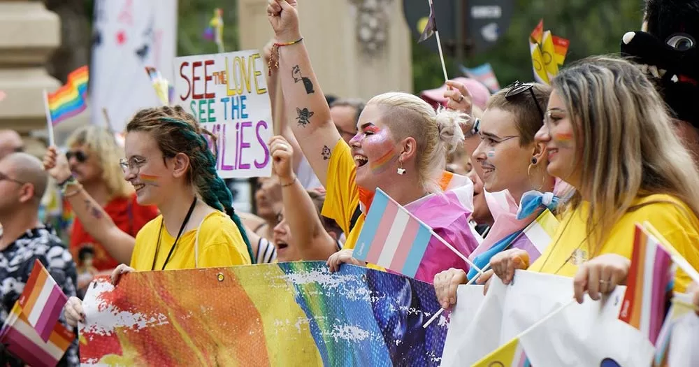 A group of people smiling and waving trans, lesbian, and rainbow Pride flags marking national coming out day