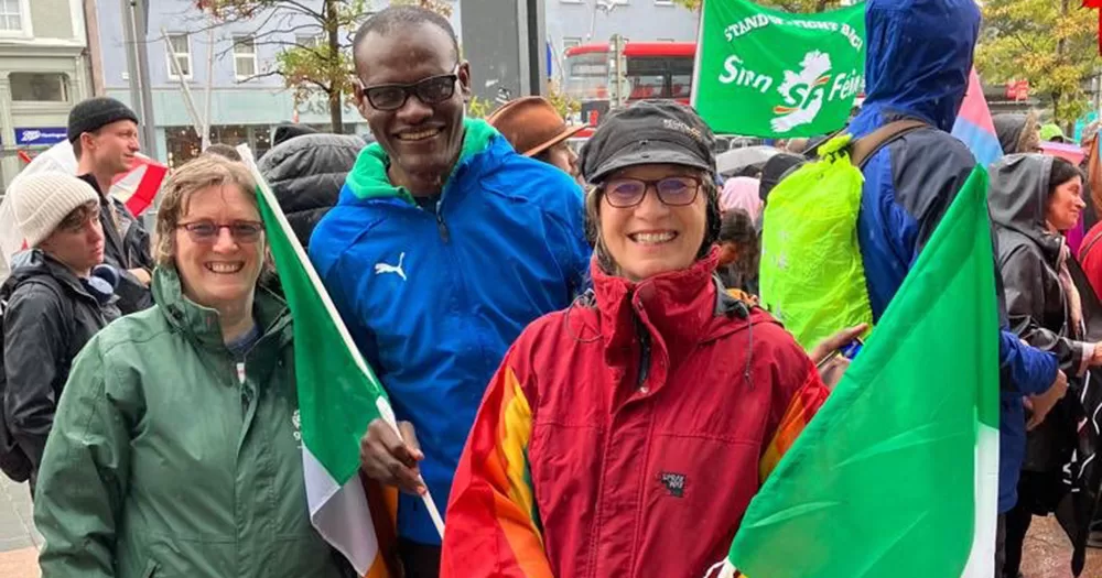 Three people at the rally in support of Cork librarians. The person on the left wears a green raincoat, the person in the middle wears a blue raincoat and holds an Irish falg, and the person on the right wears a red raincoat and holds an Irish flag and has a Pride flag around their shoulders.