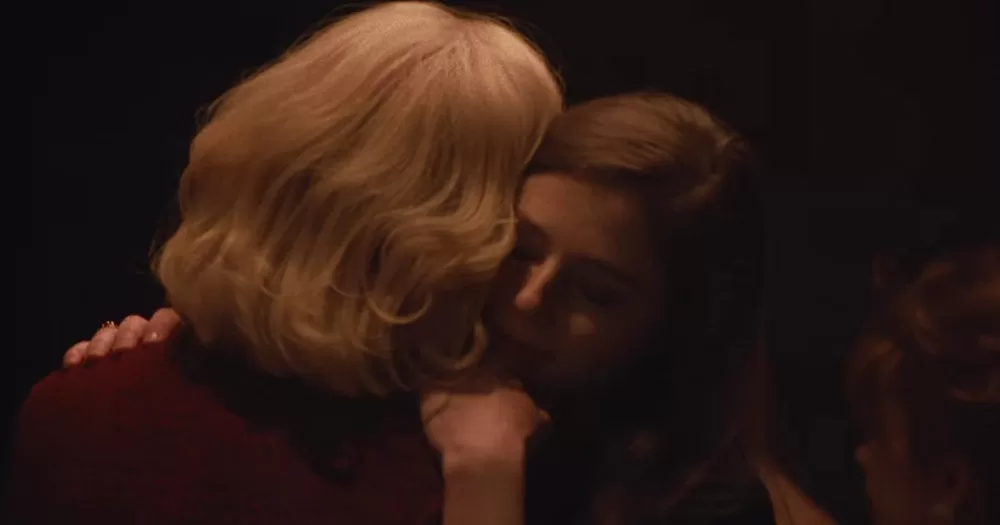 A screenshot from the Eileen trailer. It shows the back of Anne Hathaway's head, with a shoulder length bleach blonde hair cut, as she hugs Eilieen, played by Thomasin McKenzie.