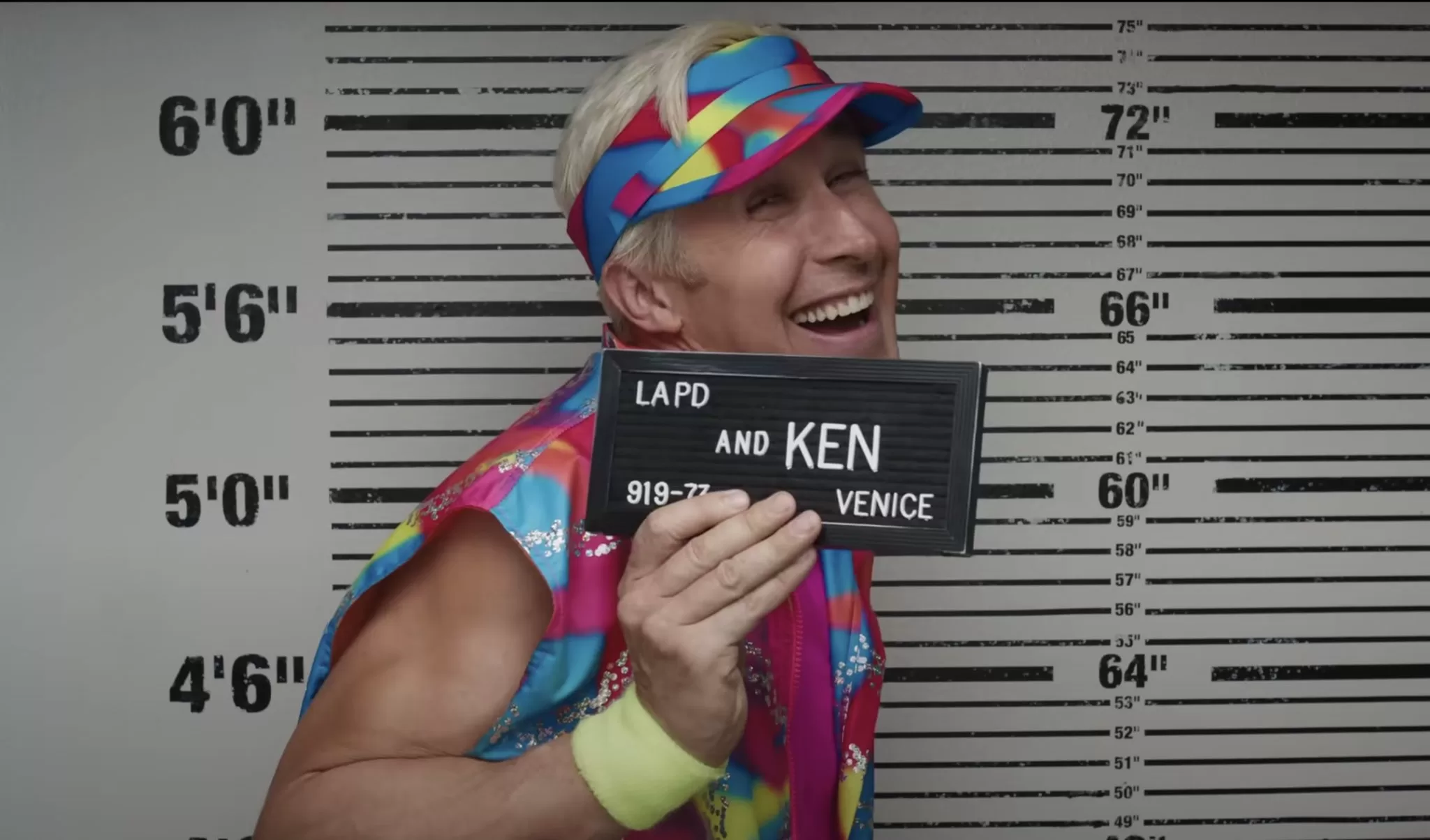 A queer Halloween costume for 2023. The image shows Ryan Gosling as Ken. The photo is from the waist up, with a man wearing a bright outfit, smiling in a mug shot and holding a sign that says LAPD and Ken.