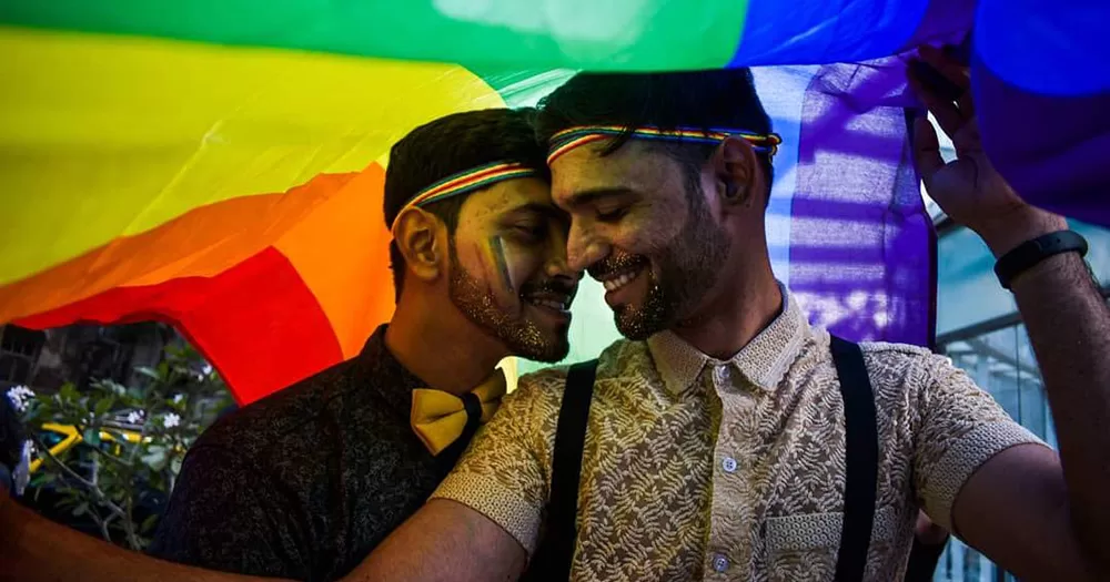 This article is about the Indian Supreme Court declining to legalise same-sex marriage. The image shows a gay Indian couple posing together while holding a rainbow flag over their heads. They have rainbow stripes painted on their faces, and are smiling looking at each other.