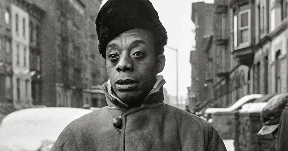 A black and white photograph of James Baldwin walking down a street. He is staring into the distance past the camera. He is wearing a black fleece hat and a suede jacket with the collar pulled up.