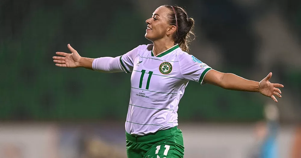 Ballon d'Or nominee Katie McCabe celebrates scoring a goal against Albania. The footballer is running and smiling with her arms spread out wide. She wears a white Ireland jersey with the number 11 on it.