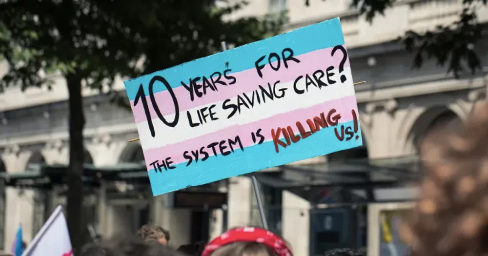 This article is about a letter to the HSE on trans healthcare. In the photo, a sign with a trans flag that reads: "10 years for life saving care? the system is killing us".