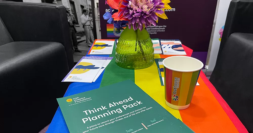 Image from LGBTQ+ Death Café. There is a rainbow coloured table, on top of which is a green pamphlet, a vase of flowers and a rainbow coloured paper cup.