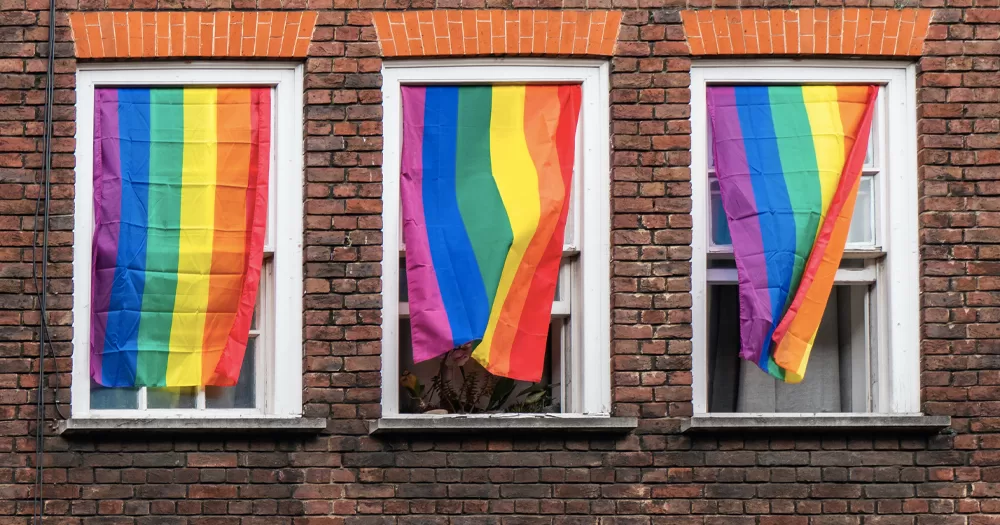 This article is about LGBTQ+ housing. In the photo, three Pride flags hanging from windows on a brick wall.