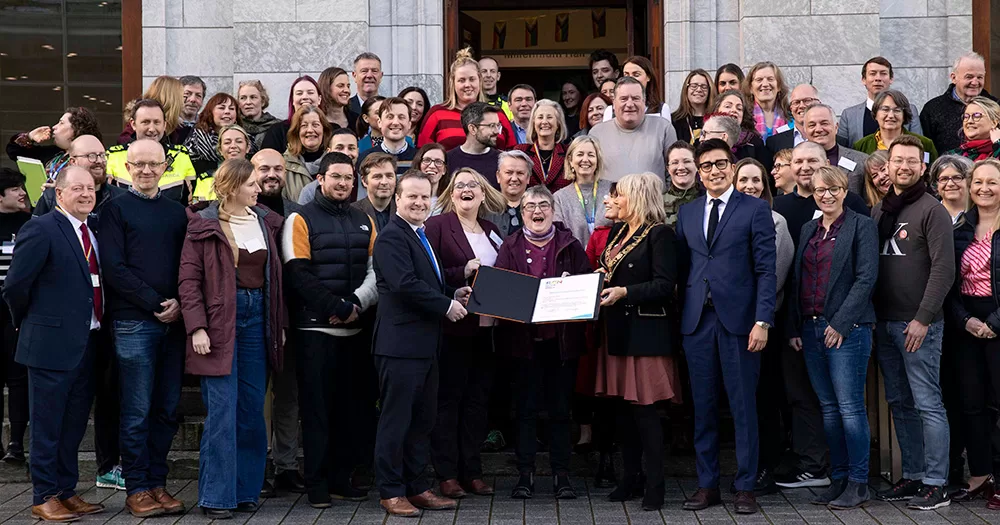 This article is about new LGBTQ+ policy guidelines by the Rainbow Cities Network. The image shows a big group of delegates of the Rainbow Cities Network posing for a photo outside City Hall, Cork. They are chatting and laughing, and holding a book open in front of them.