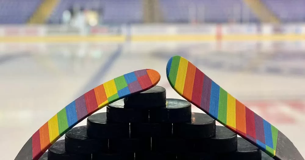 Two NHL hockey sticks wrapped in rainbow Pride tape sit atop hockey pucks with an ice rink in the background.