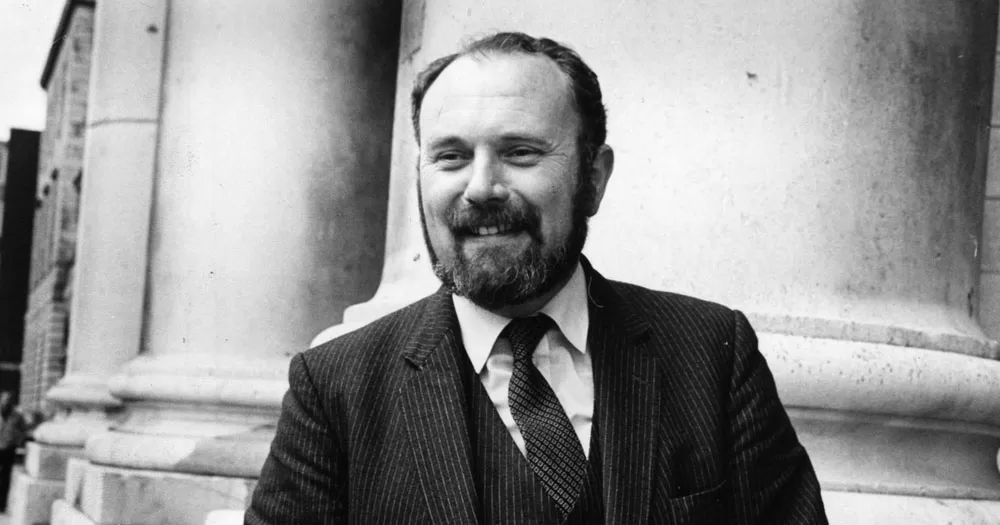 A black and white photograph shows Senator David Norris standing in front of a court building after winning his legal case. He is balding with a full beard and moustache. He is smiling and wearing a suit, tie and waistcoat.