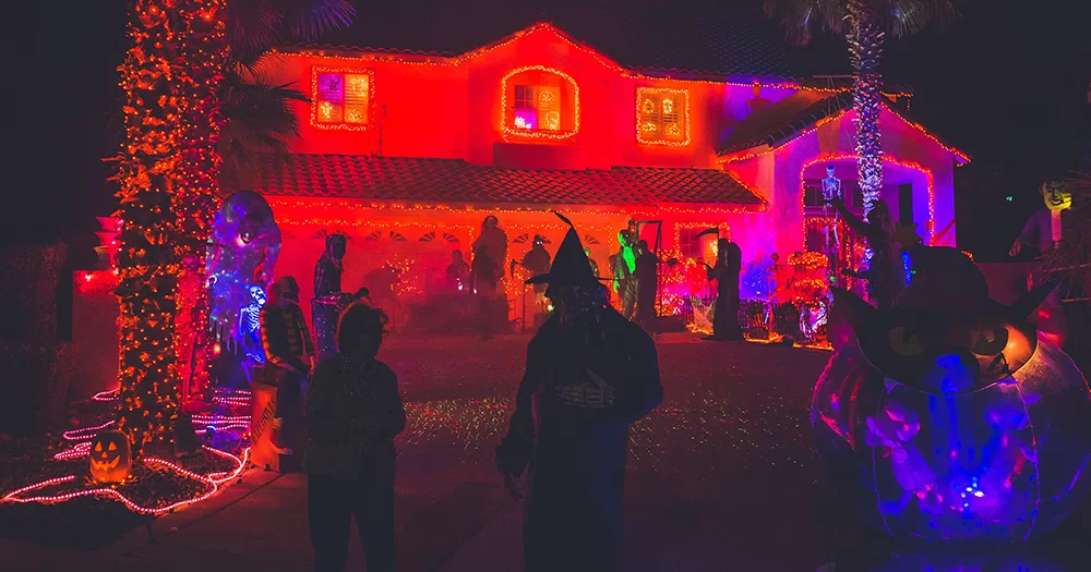 The exterior of a Halloween party. In the background there is a big house lit up in red and purple colours. There are also people outside the front of the house, with two silhouettes of people in costumes seen in focus in the foreground.