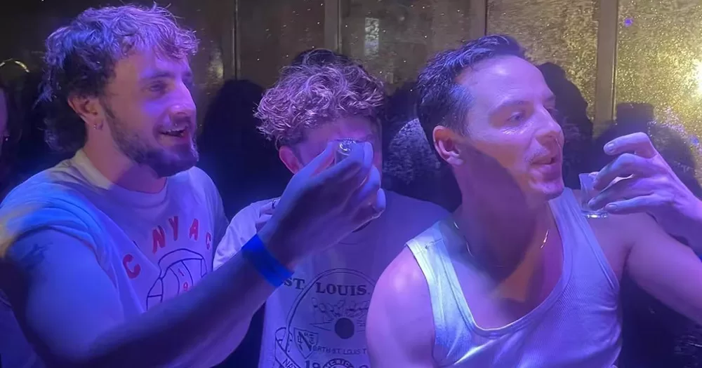 Paul Mescal, Fionn O'Shea and Andrew Scott taking shots in a gay bar. While Fionn's face is covered, Mescal and Scott are seen raising their glasses, in a crowded room with low light.