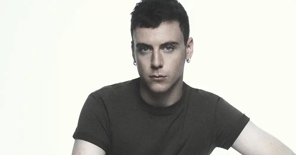 Seán McGirr photographed from the chest up. He has short black hair, small hoop earrings in both ears, and a dark coloured t-shirt. He has a serious expression and look directly down the lens.