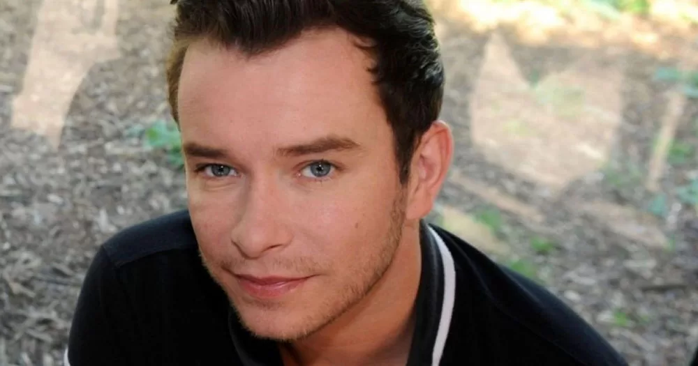 The image shows a portrait of singer Stephen Gately from Boyzone who's 14th anniversary of his death is today. In the image, he is standing in front of a window wearing a navy polo shirt with a white stripe on the collar.