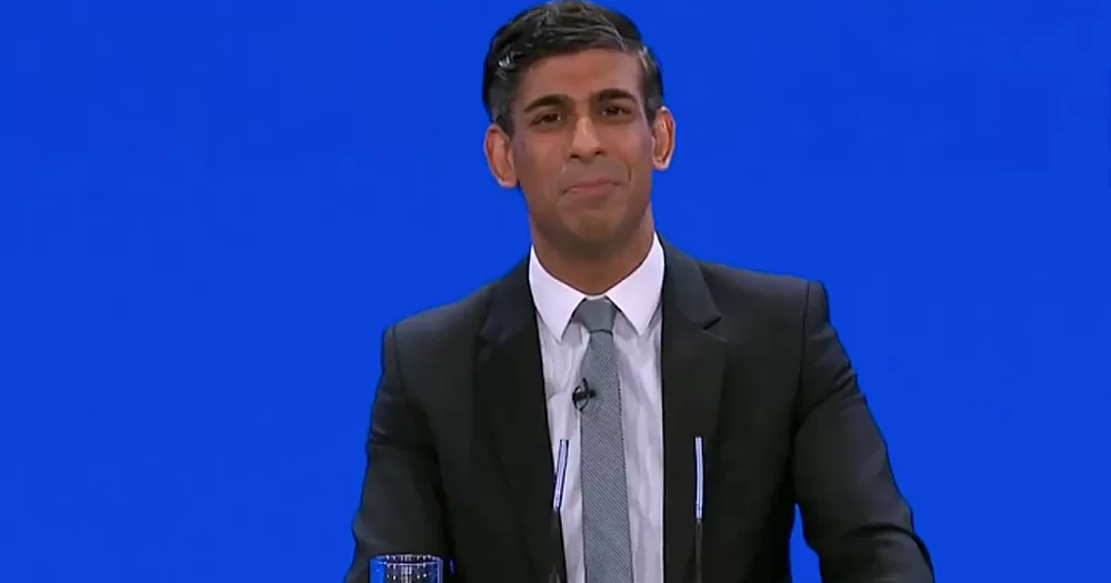 UK Prime Minister Rishi Sunak giving a speech with anti-trans views at the Tory conference, with a blue background.