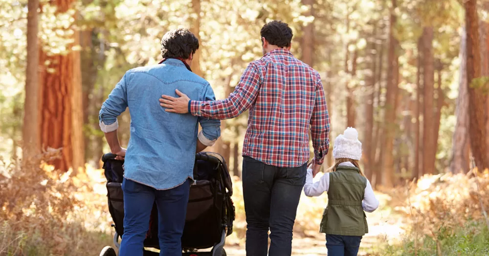 The UK government have committed to changing legislation over surrogacy laws for people living with HIV. The image shows the backs of two men walking in a forrest. One is pushing a buggy. The other man is holding the hand of a young girl and has his other arm resting on the other's back.