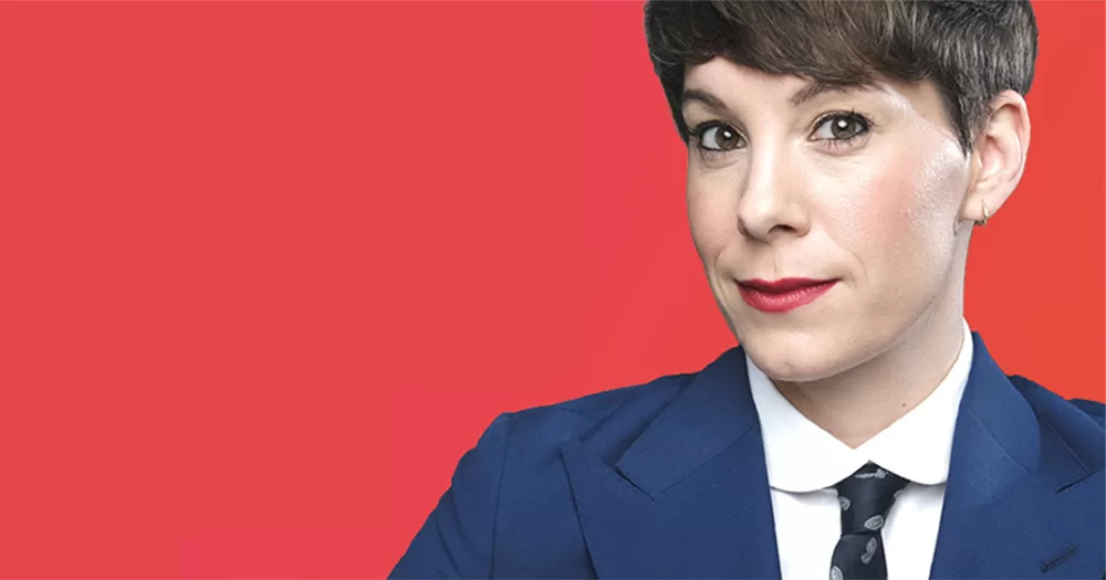 Close up of Suzi Ruffell. The comedian is show in front of a red background from the shoulders up on the right hand side of the screen, wearing a navy suit and tie with white shirt. She is smiling with red lipstick and has short brown hair.