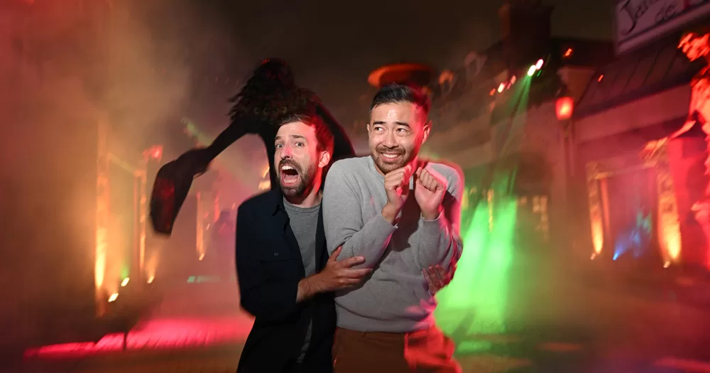 Two man hugging each other at Universal Halloween Horror Nights looking spooked at excited. In the background the silhouette of a giant spider and haunted houses