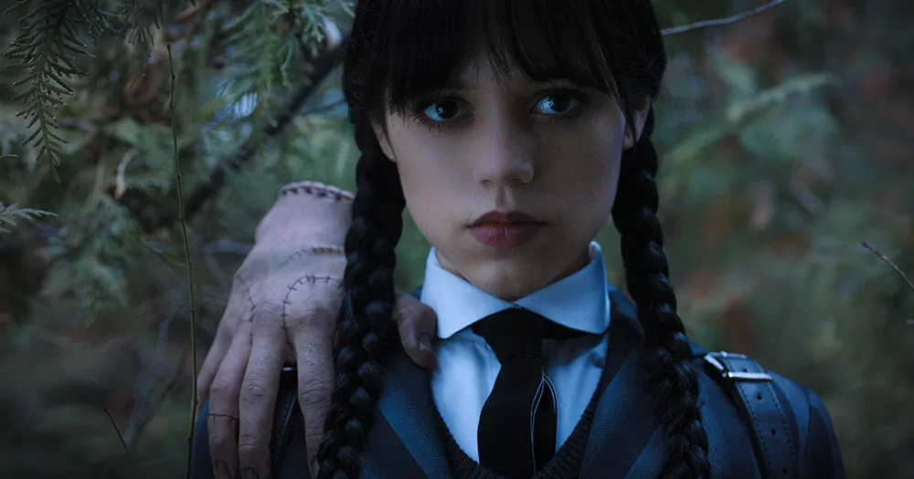 A queer Halloween costume for 2023. The image shows Jenna Ortega as Wednesday Addams. The photo is from the shoulders up, with a girl wearing a school uniform, long black plaits and a fringe. There is a severed hand perched on her shoulder.