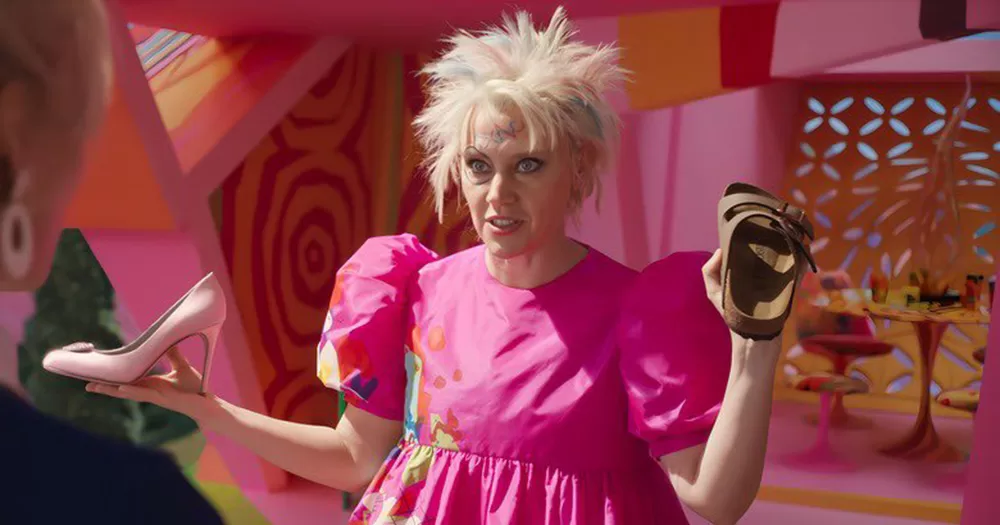 A queer costume for Halloween 2023. The image shows Kate McKinnon as Weird Barbie. She has messy blonde hair, a pink puffy dress and a heeled shoe in one hand, and a birkenstock sandal in the other.