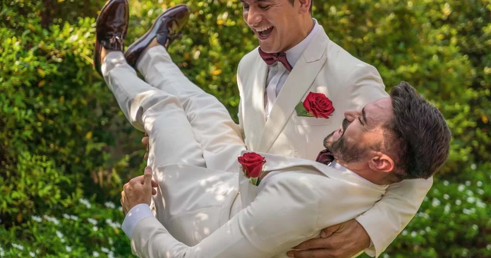 Two gay men wearing suits and playfully holding each other during garden in wedding ceremony