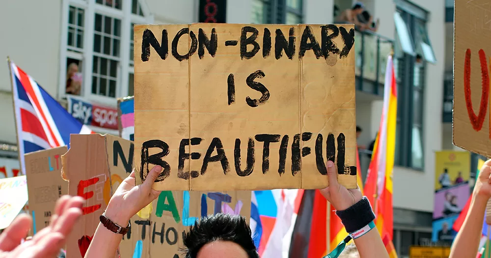 This article is about the power of language when coming out. The image shows hands holding a cardboard sign, reading "NON-BINARY IS BEAUTIFUL". In the background are other signs and flags.