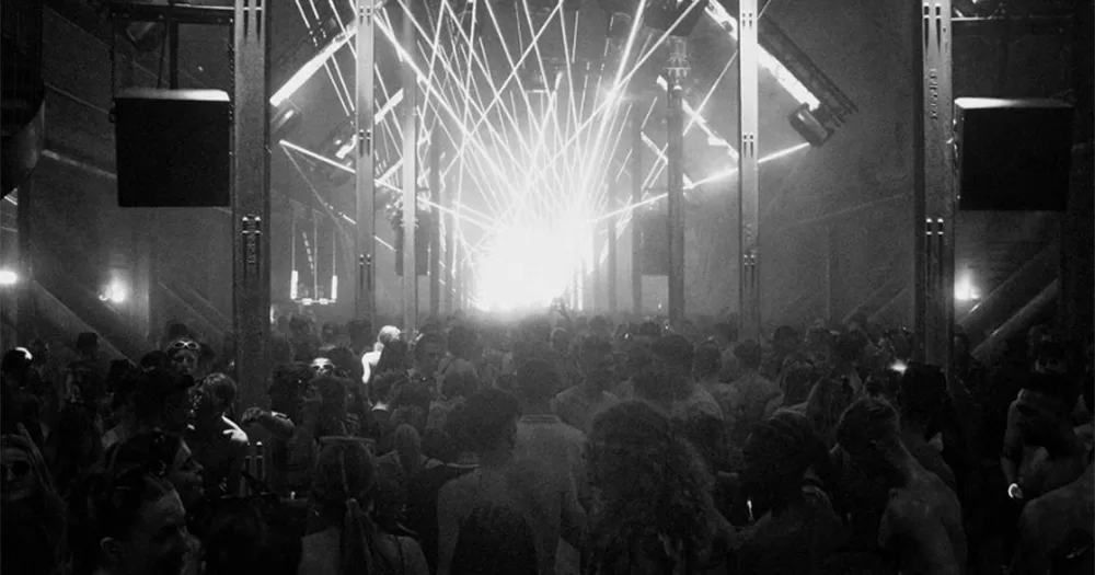 A photo from a techno rave in Cork. The image is in black and white, and is taken from the back of a large crowd at a techno party. The backs of peoples heads dancing can be seen, and strobe lights at the back of the venue where the DJs are performing are also visible.