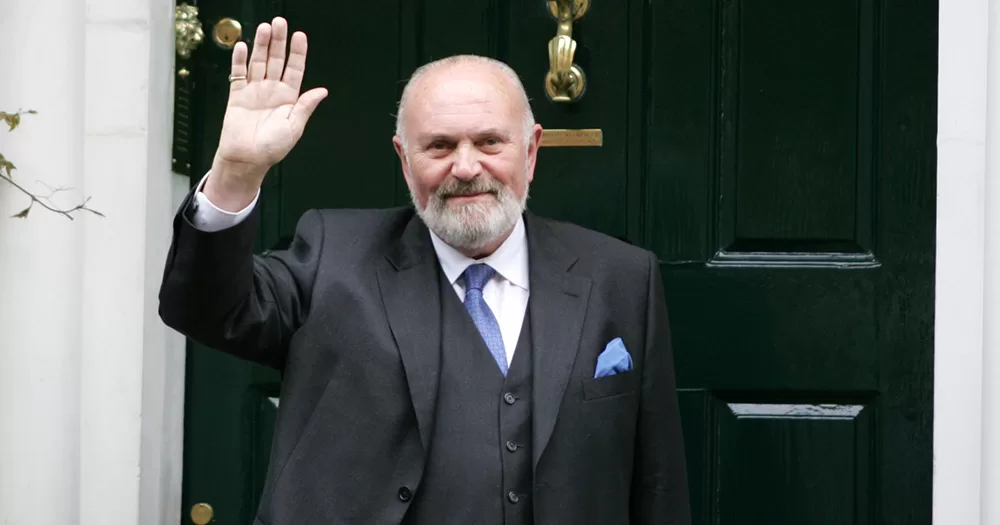Senator David Norris who has announced his retirement. Norris is photographed from the stomach up, wearing a charcoal suit, white shirt and blue tie. He stands in front of a green door, smiling and waving at the camera.