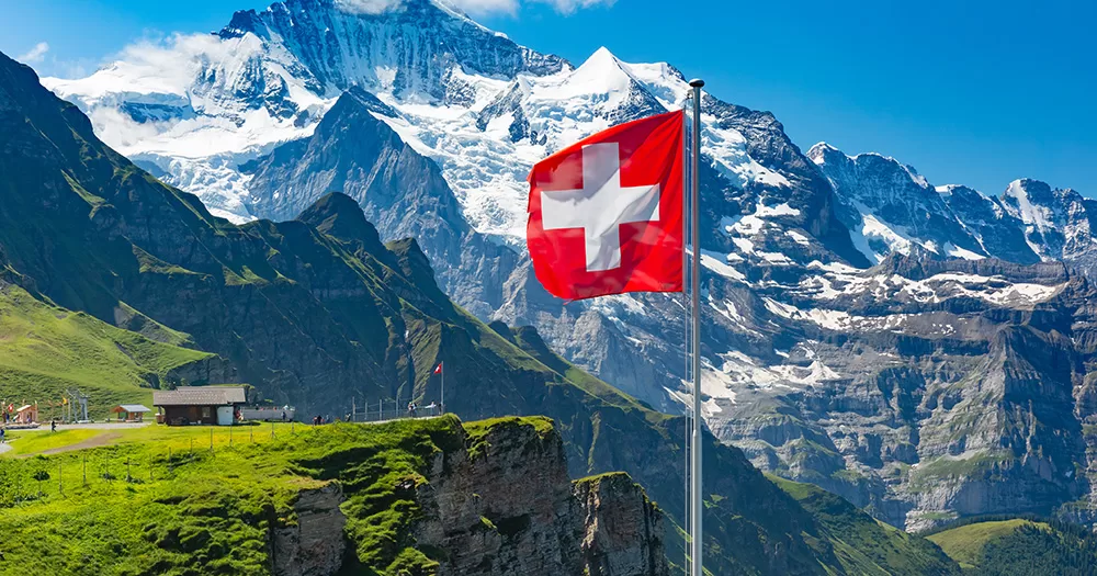 Switzerland has removed the ban on gbMSM donating blood. The image shows a Swiss flag waving and tourists admire the peaks of Monch and Jungfrau mountains on a Mannlichen viewpoint, Bernese Oberland Switzerland