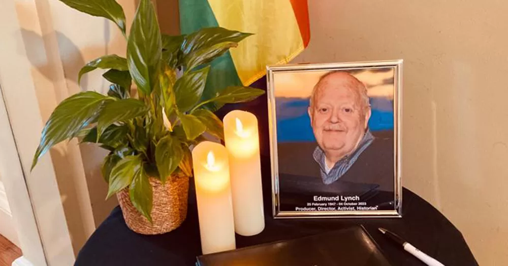 A photograph of Edmund Lynch is placed on a round wooden table with two candles, a plant and a Pride flag also displayed with it.