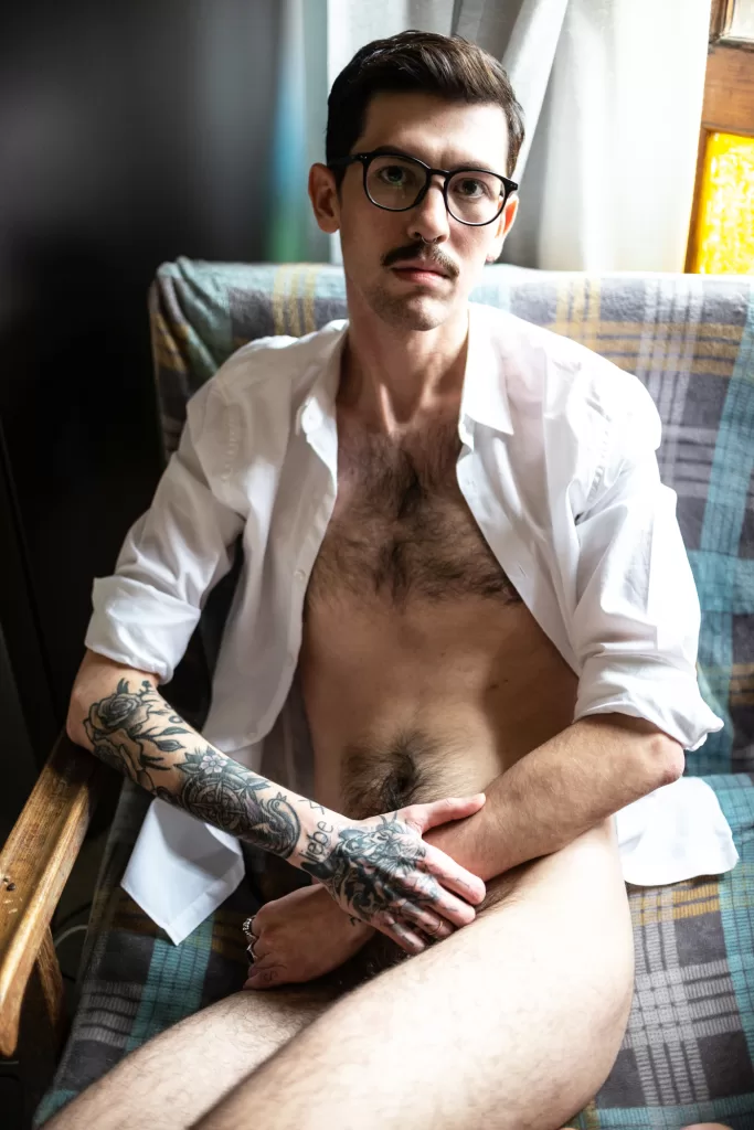 A portrait for Elska Tbilisi. A man is photographed wearing only an open white shirt and glasses. He sits on a chair with his tattooed arms in his lap, covering his groin area.