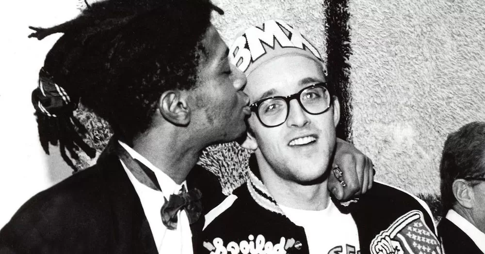 The image shows a black and white photograph of New York artists Keith Haring and Jean-Michel Basquiat. In the image, Basquiat (left), is kissing the forehead of Haring (right). Haring is wearing glasses and fez style hat with the letters BMX on it.