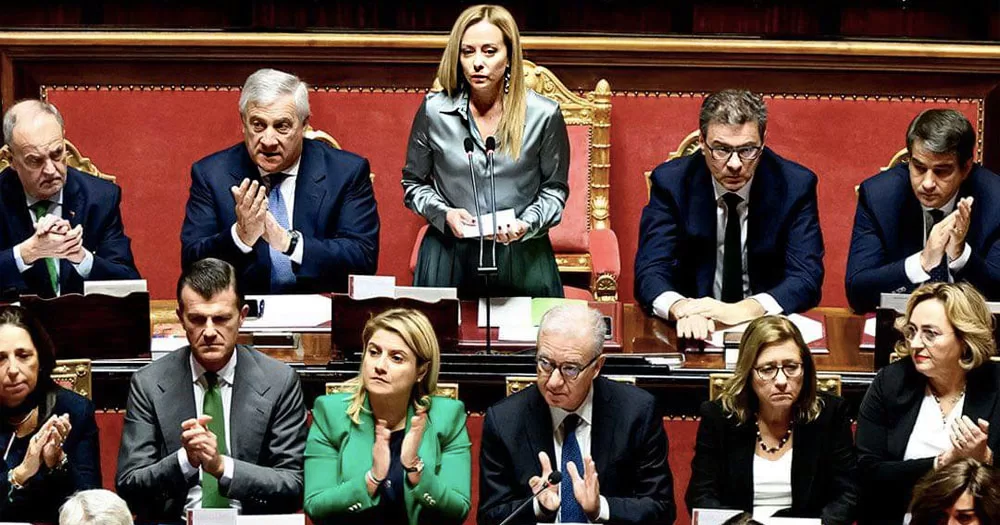 Image of far-right prime minister of Italy, Giorgia Meloni, standing to deliver a speech surrounded by her ministers, some of whom will present at a conference in Kilkenny. She is wearing a silver coloured dress, has long blonde hair and is holding paper in her hands.