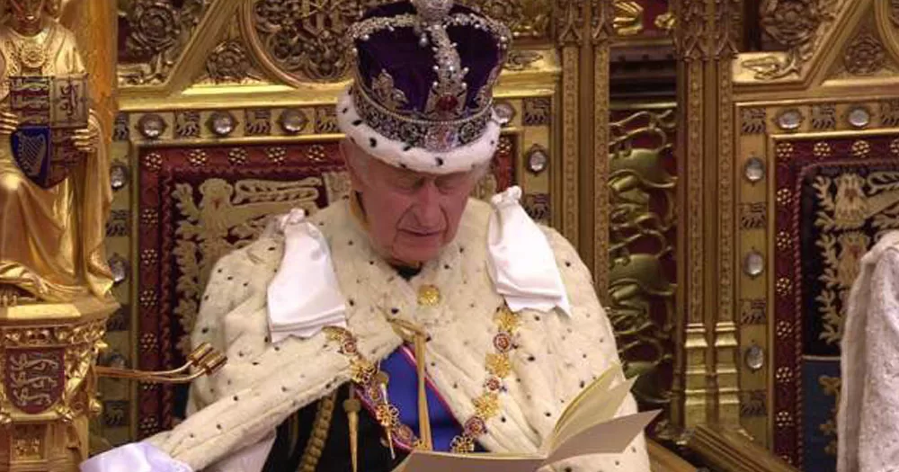 The photo shows King Charles with his head bowed as he reads the King's Speech to Parliament. He is wearing full ceremonial dress, including the crown and is sitting on a gold painted throne with embroidered upholstery featuring a repetitive pattern of lions.
