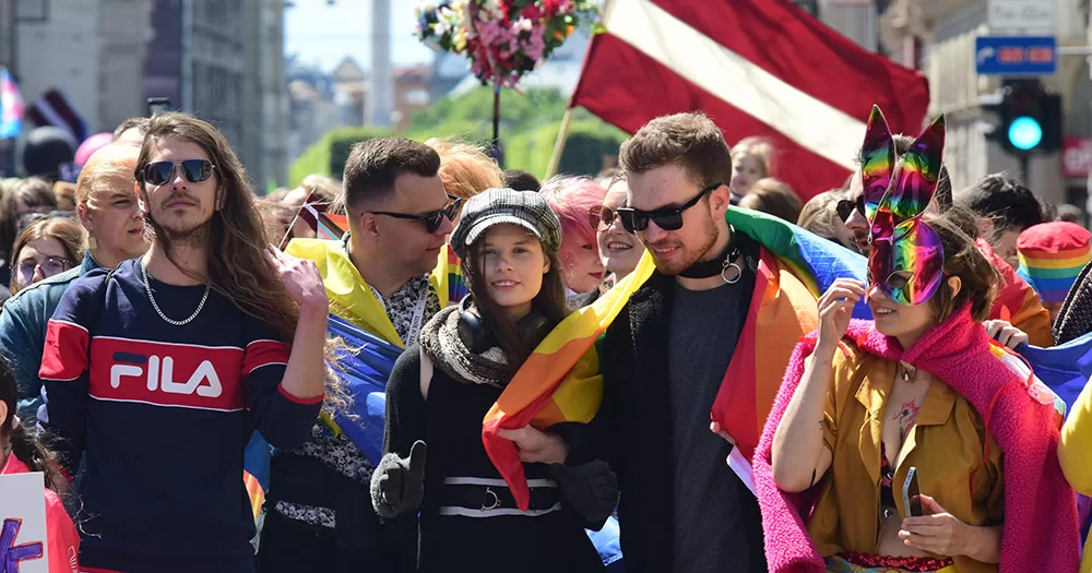 Pride being celebrated in Latvia, where same-sex civil partnerships have just been legalised. The image shows a group of participants in the Pride parade. Rainbow colours and flags are visible, as is the Latvian flag in the background.