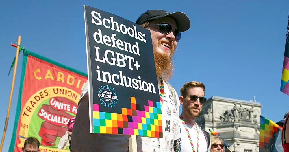 A man at a protest holds a sign reading: "Schools: defend LGBTQ+ inclusion". He can be seen smiling, wearing a black cap and sunglasses, with a blue sky in the background.