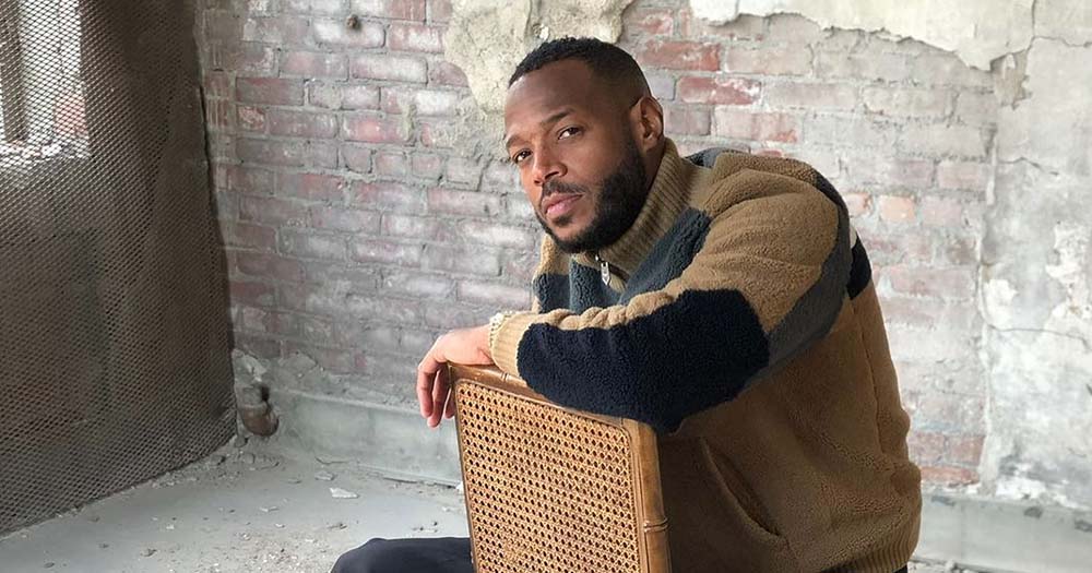 Portrait of Marlon Wayans sitting in a chair and leaning forward in front of a brick wall