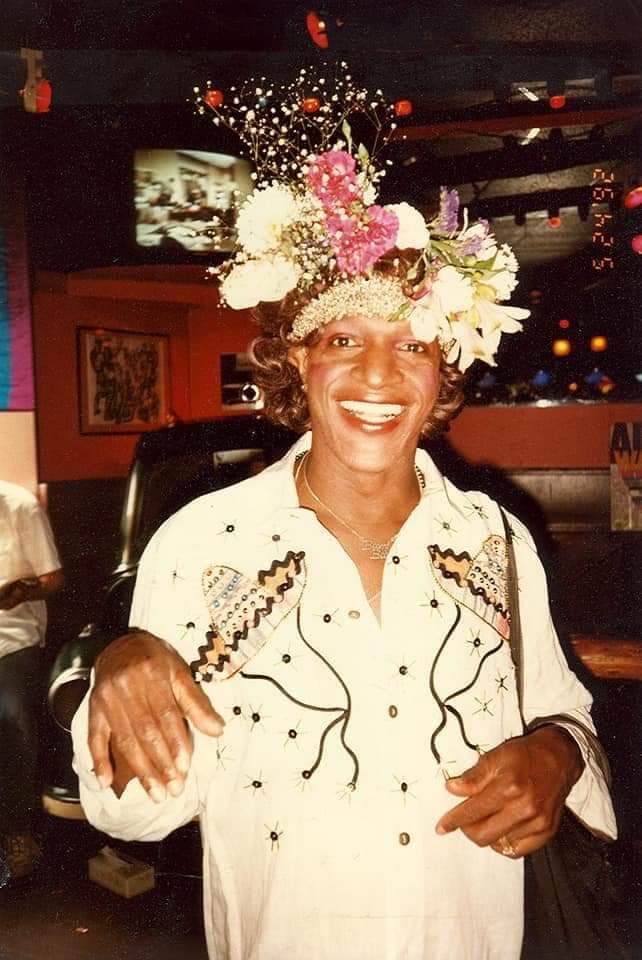 Photograph of Masha P Johnson wearing a white shirt and flowers on her hat