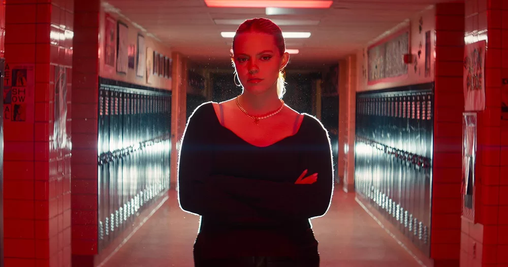 Renee Rapp as Regina George in the new Mean Girls. She is photographed in a high school hallway in red lighting with her arms crossed. She has a straight expression and looks straight down the camera.