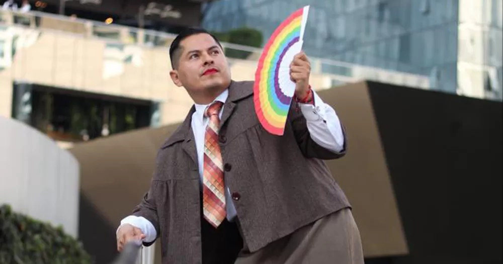 An image of Mexico non-binary activist Jesús Ociel Baena. They are photographed from the waist up, wearing a feminine suit with a shirt and tie, and holding a rainbow fan. They look off into the distance and wear red lipstick.