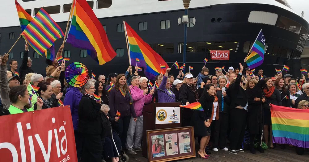 The image shows a crowd of people waving rainbow flags in front of the docked Olivia cruise boat.