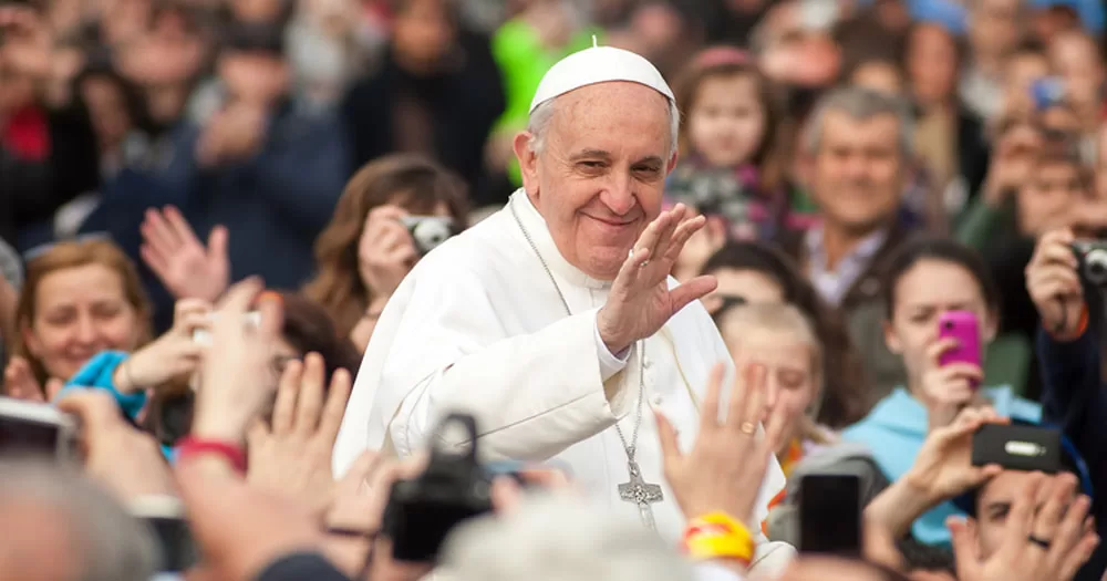 The Pope has invited a group of trans women to a meal for World Day of Poor. The image shows Pope Francis standing above a crowd waving. He is wearing traditional white robes and a white head covering. He is smiling.