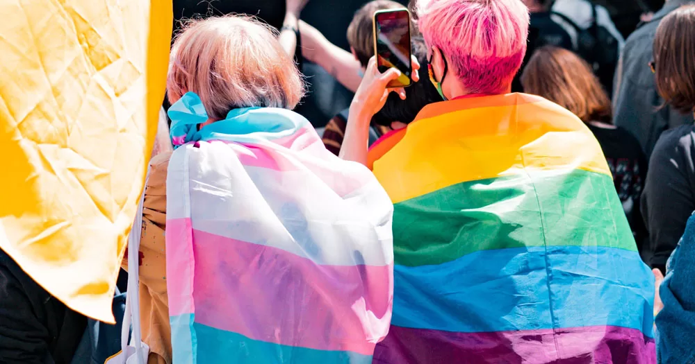 The image shows two people who have come out about their LGBTQ+ identity at a Pride event. They are standing with their backs to the camera. One has a trans Pride flag wrapped around their shoulders and one has a rainbow Pride flag wrapped around their shoulders.