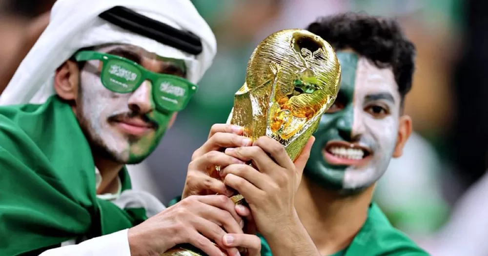 Saudi Arabia fans holding a World Cup trophy. Both men have their faces painted green and yellow, with one wearing green sunglasses as well. They hold the gold trophy in front of them as they pose for the camera.