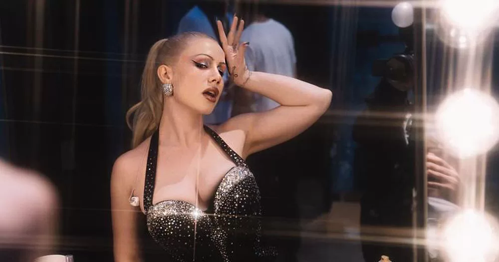 The image shows a portrait of singer Slayyyter. She is sitting in front of a dressing room mirror surrounded with light bulbs. She is wearing a black halter neck bodice with diamontés across the cleavage. She has the back of one hand raised to her forehead in a fainting pose.